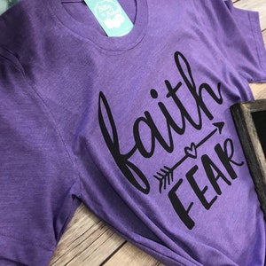 May include: A purple t-shirt with the words "faith" above "fear" in black script and an arrow design. The shirt has a round neckline and a tag that says "Bitty & Boho". The shirt is on a wooden surface.