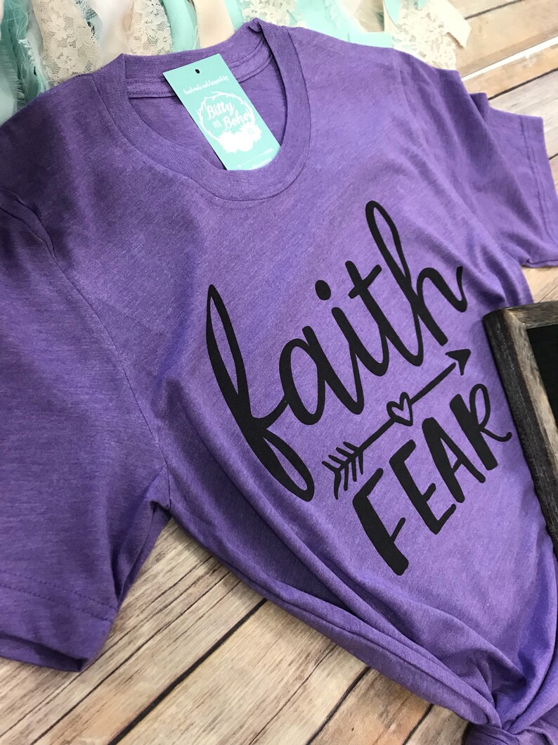 May include: A purple t-shirt with the words "faith" above "fear" in black script and an arrow design. The shirt has a round neckline and a tag that says "Bitty & Boho". The shirt is on a wooden surface.