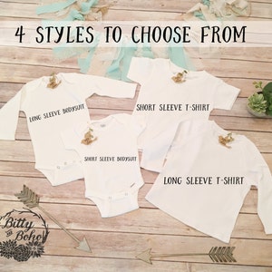 K&ouml;nnte beinhalten: Vier wei&szlig;e Babykleidungsst&uuml;cke: ein lang&auml;rmliger Body, ein kurz&auml;rmliger Body, ein kurz&auml;rmliges T-Shirt und ein lang&auml;rmliges T-Shirt. Der Text "4 STYLES TO CHOOSE FROM" steht oben. Das Bitty and Boho-Logo befindet sich unten.