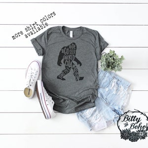 Puede incluir: Una camiseta gris oscuro con un gr&aacute;fico de Bigfoot que dice "Hide and Seek World Champion Since 1969". La camiseta se combina con zapatillas blancas y pantalones cortos de mezclilla. El texto en la imagen dice "more shirt colors available."