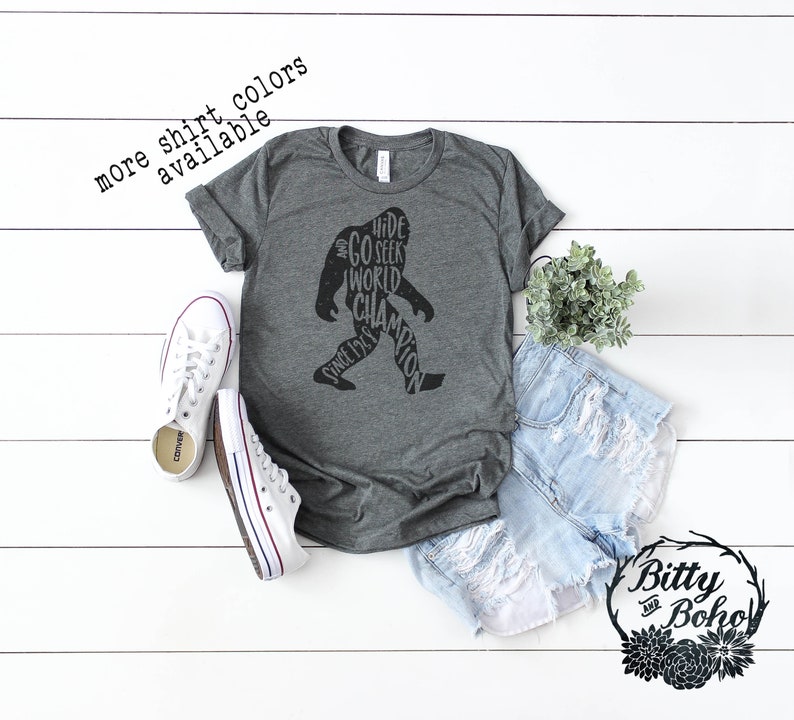 Puede incluir: Una camiseta gris oscuro con un gr&aacute;fico de Bigfoot que dice "Hide and Seek World Champion Since 1969". La camiseta se combina con zapatillas blancas y pantalones cortos de mezclilla. El texto en la imagen dice "more shirt colors available."
