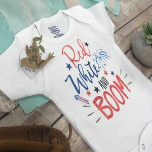 May include: A white short-sleeved bodysuit featuring the text Red White and BOOM in red and blue lettering. The design includes graphic illustrations of a firework, stars, and a rocket. A brown bird-shaped tag with the text Bitty Boho is attached.