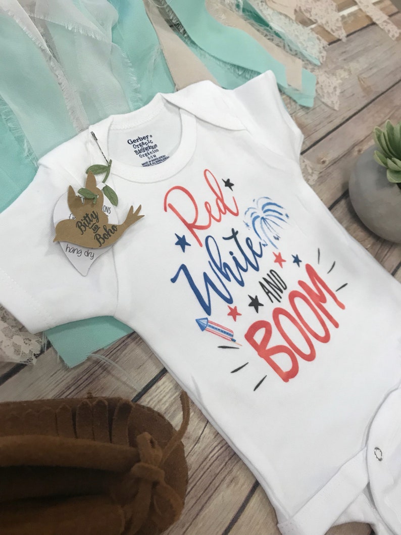 May include: A white short-sleeved bodysuit featuring the text Red White and BOOM in red and blue lettering. The design includes graphic illustrations of a firework, stars, and a rocket. A brown bird-shaped tag with the text Bitty Boho is attached.