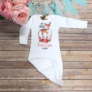 Puede incluir: Bata de bebé blanca con mangas largas. La bata presenta un gráfico de ciervo con el texto "Oh Deer, Sadie Lynn is here" en rosa. La bata está decorada con detalles florales y está hecha de tela suave.