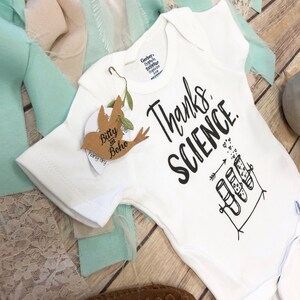 May include: A white baby grow with black text that reads "Thanks, Science." The grow features a drawing of three test tubes with hearts inside. The test tubes are on a stand with an arrow pointing to them. There are small hearts scattered around the test tubes.