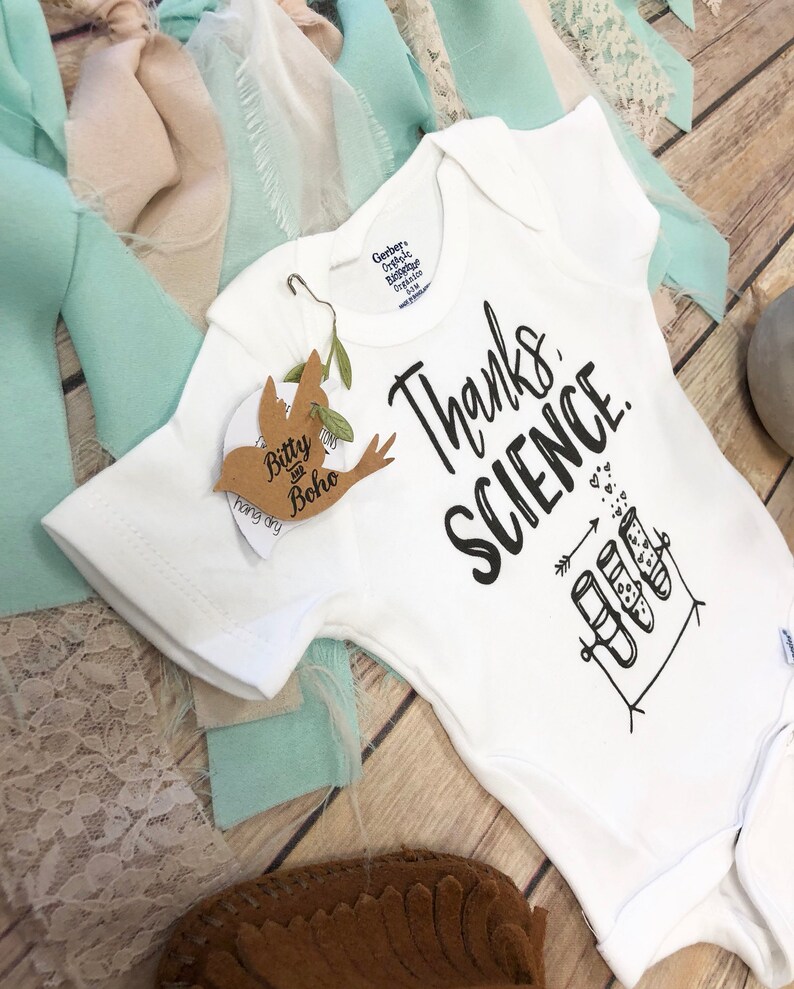 May include: A white baby grow with black text that reads "Thanks, Science." The grow features a drawing of three test tubes with hearts inside. The test tubes are on a stand with an arrow pointing to them. There are small hearts scattered around the test tubes.