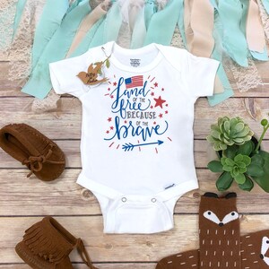 K&ouml;nnte beinhalten: Wei&szlig;er Baby-Body mit dem Text "Land of the Free Because of the Brave" in Blau und Rot, mit einer amerikanischen Flagge. Enth&auml;lt braune Mokassins und Socken mit Fuchsmotiv. Dekorative B&auml;nder und eine kleine Pflanze sind ebenfalls vorhanden.