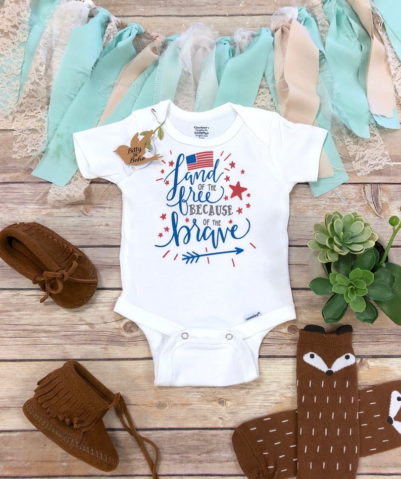 K&ouml;nnte beinhalten: Wei&szlig;er Baby-Body mit dem Text "Land of the Free Because of the Brave" in Blau und Rot, mit einer amerikanischen Flagge. Enth&auml;lt braune Mokassins und Socken mit Fuchsmotiv. Dekorative B&auml;nder und eine kleine Pflanze sind ebenfalls vorhanden.