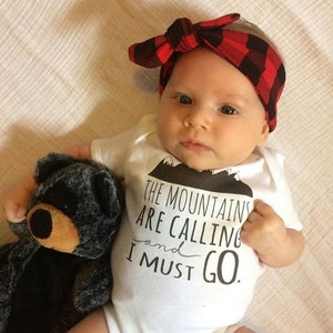 May include: A white baby onesie with black text that reads "The mountains are calling and I must go."  The onesie features a small black mountain graphic. A baby is wearing the onesie and holding a gray teddy bear. The baby is wearing a red and black plaid headband.