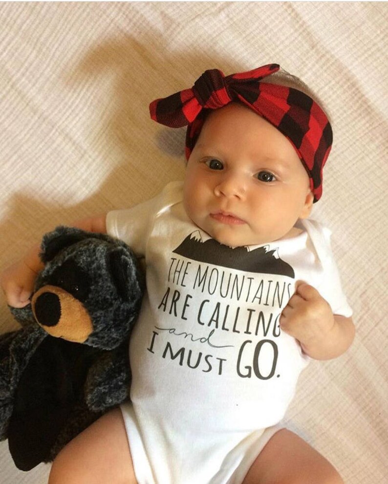 May include: A white baby onesie with black text that reads "The mountains are calling and I must go."  The onesie features a small black mountain graphic. A baby is wearing the onesie and holding a gray teddy bear. The baby is wearing a red and black plaid headband.