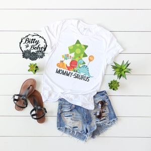 May include: White t-shirt with a green, yellow, and blue dinosaur graphic. The dinosaurs are holding a sign that says "Dino Mite!" and a balloon that says "One". The text "Mommy-saurus" is printed below the dinosaurs.