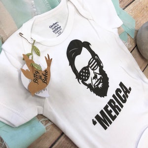 Puede incluir: Un body blanco de manga corta para beb&eacute; con un gr&aacute;fico negro de una persona con barba que lleva gafas de sol con un patr&oacute;n de estrellas. El texto 'MERICA. est&aacute; impreso en negro debajo del gr&aacute;fico. Una etiqueta marr&oacute;n Bitty and Boho est&aacute; sujeta.