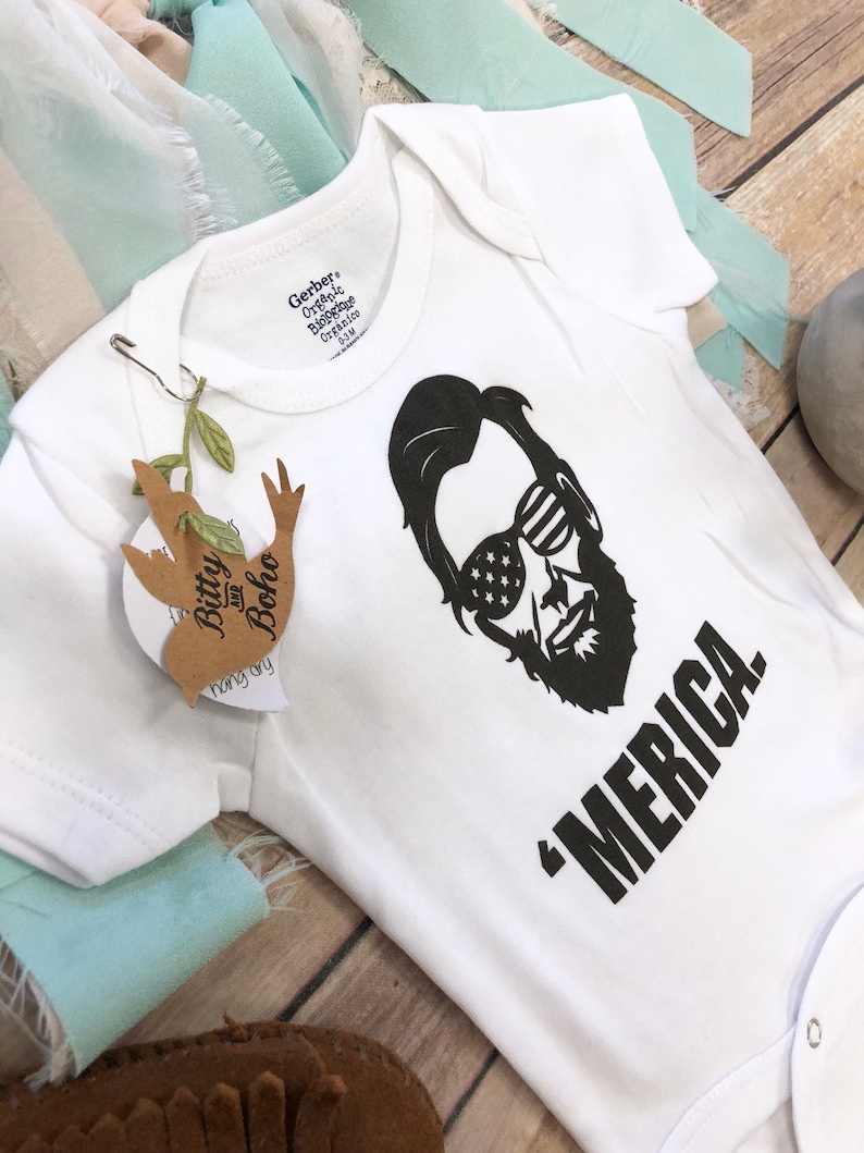 Puede incluir: Un body blanco de manga corta para beb&eacute; con un gr&aacute;fico negro de una persona con barba que lleva gafas de sol con un patr&oacute;n de estrellas. El texto 'MERICA. est&aacute; impreso en negro debajo del gr&aacute;fico. Una etiqueta marr&oacute;n Bitty and Boho est&aacute; sujeta.
