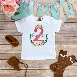 K&ouml;nnte beinhalten: Wei&szlig;es T-Shirt mit einer roten Zahl 2, die von einem Blumenkranz umgeben ist. Der Name "Caroline" ist in Kursivschrift &uuml;ber der Zahl geschrieben. Das Shirt liegt auf einem Holzgrund mit braunen Mokassins und braunen Fuchs-Socken.