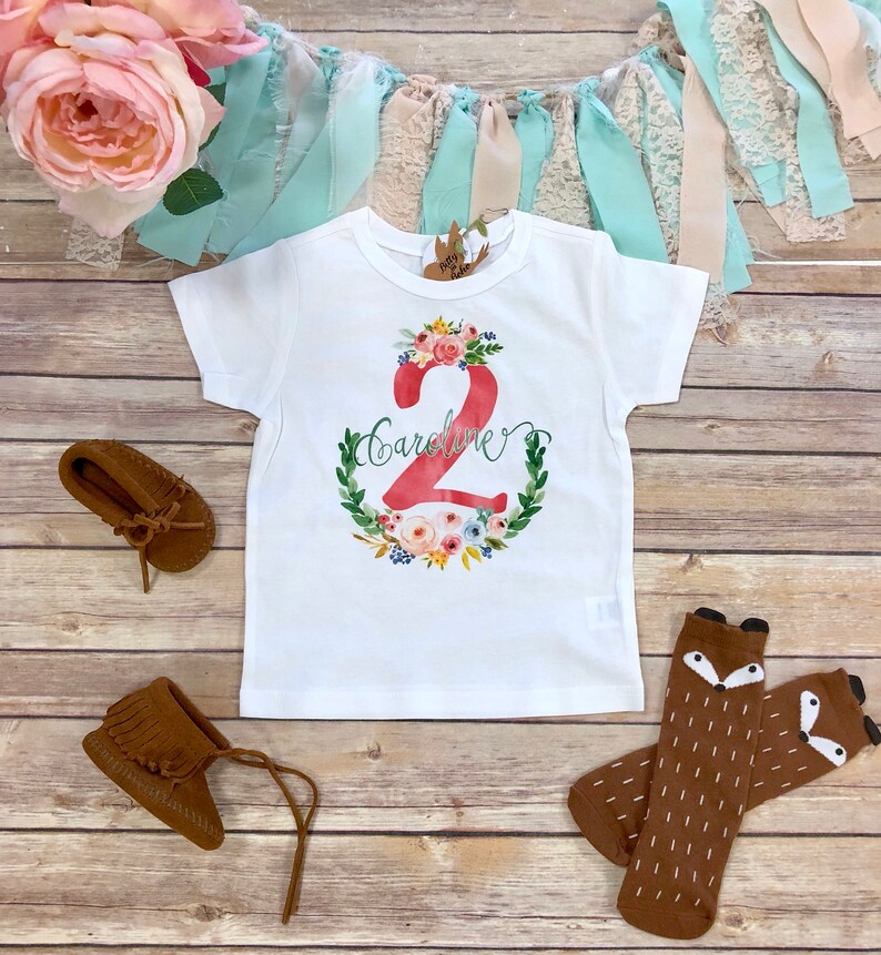 K&ouml;nnte beinhalten: Wei&szlig;es T-Shirt mit einer roten Zahl 2, die von einem Blumenkranz umgeben ist. Der Name "Caroline" ist in Kursivschrift &uuml;ber der Zahl geschrieben. Das Shirt liegt auf einem Holzgrund mit braunen Mokassins und braunen Fuchs-Socken.