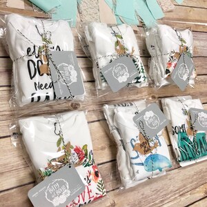 May include: Several white baby onesies, each packaged in clear plastic and tied with twine. Each onesie has a unique design, including text and floral or animal graphics. Gray tags with the "Bitty and Boho" logo are attached.