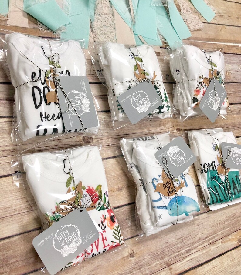May include: Several white baby onesies, each packaged in clear plastic and tied with twine. Each onesie has a unique design, including text and floral or animal graphics. Gray tags with the "Bitty and Boho" logo are attached.