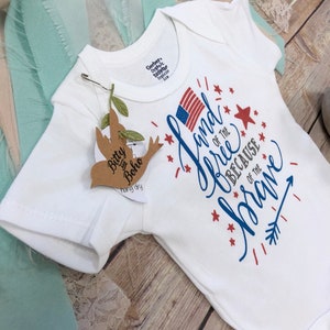 K&ouml;nnte beinhalten: Wei&szlig;er Baby-Body mit dem Text "Land of the Free Because of the Brave" in Rot und Blau, mit einer amerikanischen Flagge. Ein kleines Etikett mit den Worten "Bitty Boho" ist angebracht. Der Body ist f&uuml;r 6-9 Monate.