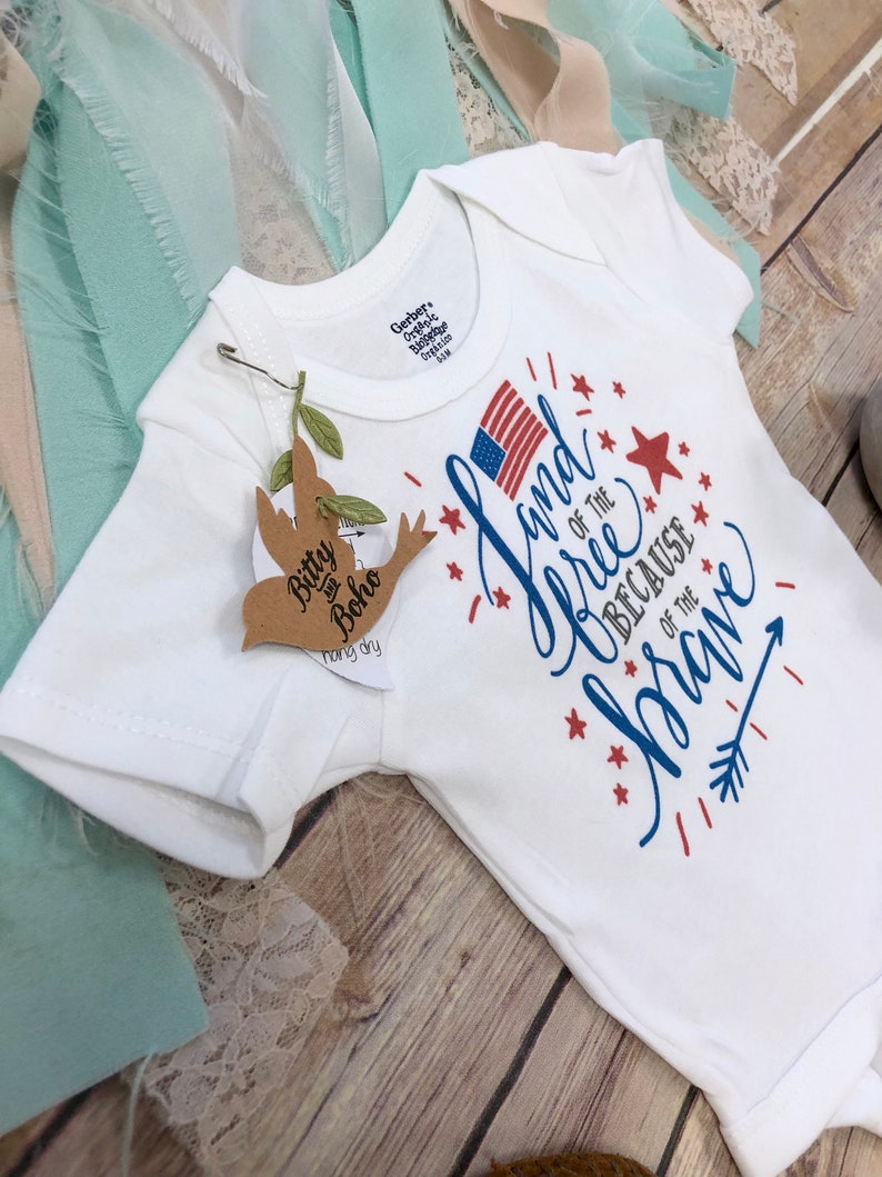 K&ouml;nnte beinhalten: Wei&szlig;er Baby-Body mit dem Text "Land of the Free Because of the Brave" in Rot und Blau, mit einer amerikanischen Flagge. Ein kleines Etikett mit den Worten "Bitty Boho" ist angebracht. Der Body ist f&uuml;r 6-9 Monate.