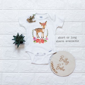 May include: White baby onesie with a watercolor illustration of a deer wearing a floral crown, surrounded by flowers and greenery. The text "short or long sleeve available" is printed on the right side. A wooden disc with the text "Bitty + Boho" is also present.