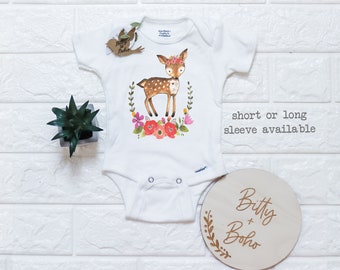 May include: White baby onesie with a watercolor illustration of a deer wearing a floral crown, surrounded by flowers and greenery. The text "short or long sleeve available" is printed on the right side. A wooden disc with the text "Bitty + Boho" is also present.