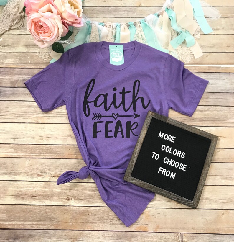 May include: A purple t-shirt with the words "faith" above "fear" in black script, with an arrow and heart design. A small sign reads "More colors to choose from."