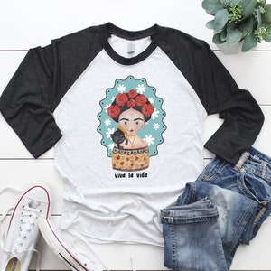 以下が含まれることがあります： グレーと白のラグランスリーブTシャツ。フリーダ・カーロ風のイラストと黒猫が描かれています。デザインには、白い花と「viva la vida」の文字が入ったターコイズ色のボーダーが含まれています。