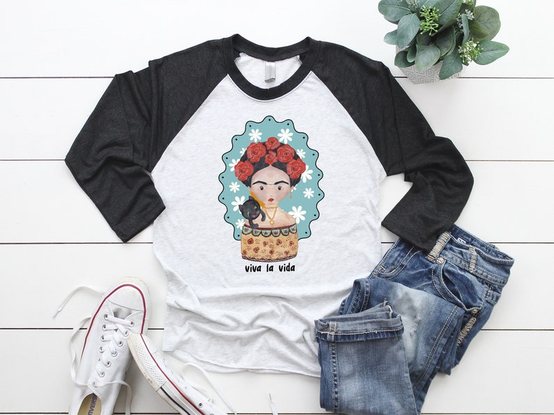 以下が含まれることがあります： グレーと白のラグランスリーブTシャツ。フリーダ・カーロ風のイラストと黒猫が描かれています。デザインには、白い花と「viva la vida」の文字が入ったターコイズ色のボーダーが含まれています。