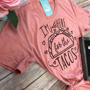 May include: A pink t-shirt with the text "I'm just here for the tacos" in black lettering with a taco graphic and hearts.
