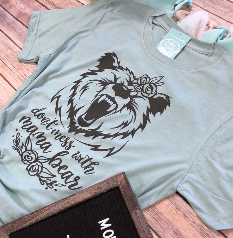 May include: A light blue t-shirt with a black graphic of a bear's head with the text "don't mess with mama bear". The bear has an open mouth and a floral crown. The shirt has a tag that says "Betty & Boho".
