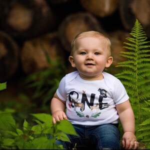 Op de afbeelding: Een baby in een witte romper met het woord "ONE" op de voorkant. Het woord "ONE" is versierd met dierenillustraties. De baby zit buiten met groen gebladerte en een achtergrond van boomstammen.