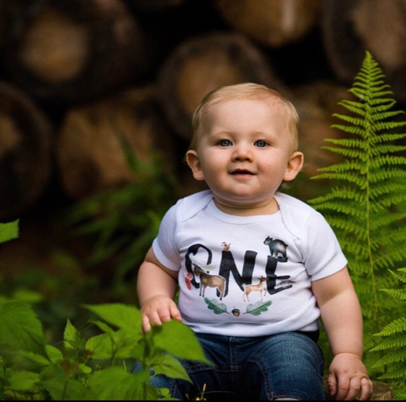 Op de afbeelding: Een baby in een witte romper met het woord "ONE" op de voorkant. Het woord "ONE" is versierd met dierenillustraties. De baby zit buiten met groen gebladerte en een achtergrond van boomstammen.
