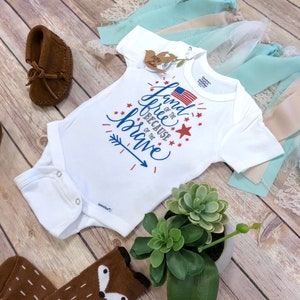 K&ouml;nnte beinhalten: Wei&szlig;er Baby-Body mit dem Text "Land of the Free Because of the Brave" in Blau und Rot, mit einer amerikanischen Flagge und Sternen. Braune Mokassins und Socken mit Fuchsmotiv sind ebenfalls zu sehen.