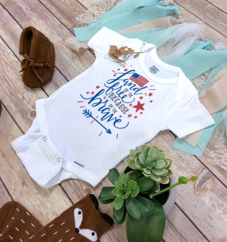 K&ouml;nnte beinhalten: Wei&szlig;er Baby-Body mit dem Text "Land of the Free Because of the Brave" in Blau und Rot, mit einer amerikanischen Flagge und Sternen. Braune Mokassins und Socken mit Fuchsmotiv sind ebenfalls zu sehen.