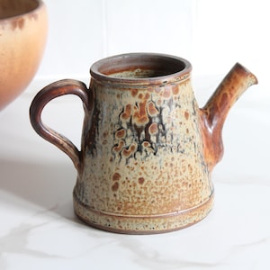 Peut inclure: Un arrosoir en céramique avec une poignée et un bec marron. Le corps du pot est beige avec des motifs mouchetés marron et noirs. Un grand bol marron est à l'arrière-plan.