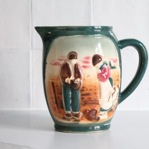 Può includere: Brocca vintage in ceramica con bordo e manico verde scuro. Il corpo presenta una scena a rilievo di due figure e un cane in un campo, con colori marrone, verde e crema. La brocca ha un beccuccio per versare.