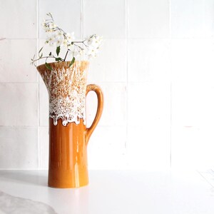 May include: Tall, orange ceramic pitcher with a textured, white and brown upper section. The pitcher has a curved handle and is filled with white flowers and green leaves. The pitcher is on a white surface against a white tiled background.