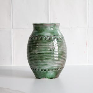 Peut inclure: Un vase en céramique vert avec un motif texturé et nervuré. Le vase a une forme arrondie avec un bord légèrement évasé et une série de petites indentations circulaires autour du col. La couleur est un vert sourd avec des touches de rose.