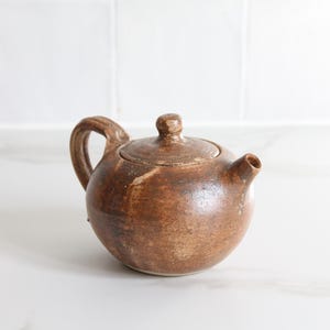 Peut inclure: Petite théière en céramique marron avec une anse incurvée et un couvercle. La théière a un bec verseur et une surface texturée, suggérant une esthétique artisanale ou rustique. Idéale pour infuser le thé.