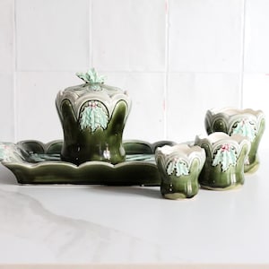 Può includere: Un set da portata in ceramica verde con un vassoio, un contenitore con coperchio e tre piccole tazze. Il set presenta un design smerlato con un motivo a foglie in bianco e verde. I pezzi sono disposti su una superficie bianca.