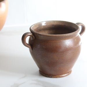 Puede incluir: Una olla de cerámica marrón con dos asas curvas. La olla tiene un cuerpo redondeado y un borde ligeramente ensanchado. El exterior tiene un acabado liso y vidriado. La olla probablemente se utiliza para cocinar o almacenar.