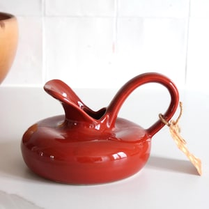 Peut inclure: Une cruche en céramique rouge brillant avec une anse incurvée et un bec verseur. La cruche a une base ronde et une étiquette attachée à l'anse. Un grand bol marron est en arrière-plan.