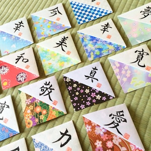 Puede incluir: Un conjunto de 14 adivinanzas de origami, cada una con un carácter japonés diferente escrito en ella. Las adivinanzas están hechas de papel estampado de colores y están dobladas en forma cuadrada.