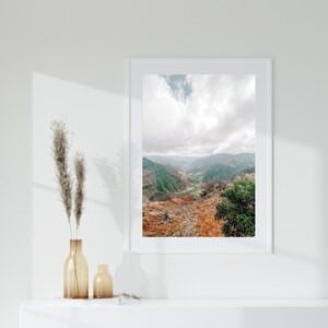 May include: A framed landscape photograph of a canyon with green and brown terrain under a cloudy sky. The photograph is in a white frame and is displayed on a white shelf with two amber glass vases.