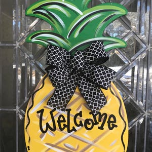 Può includere: Un appendiabiti a forma di ananas in legno, con la parte superiore verde e il corpo giallo. La parola "Welcome" è scritta in nero. Un fiocco a fantasia bianca e nera è attaccato.