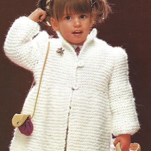 May include: A young girl wearing a white knitted cardigan with a button closure and a small brown purse. She is holding a wicker basket with a pink and white lining.