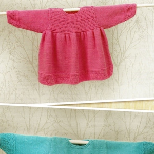 Pode incluir: Dois suéteres de malha para bebê, um rosa e outro azul turquesa, pendurados em um cabide de madeira sobre um fundo branco com um padrão de galhos de árvore.
