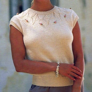 May include: A white sleeveless knitted top with a delicate lace pattern around the neckline. The neckline is adorned with small pink flowers.