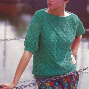 Puede incluir: Una mujer con un suéter de crochet verde y una falda de estampado floral. Está de pie frente a una valla de cadena.