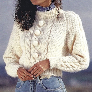 Könnte beinhalten: Ein weißer Strickpullover mit Zopfmuster und Bommelknöpfen. Der Pullover hat einen hohen Kragen und lange Ärmel.